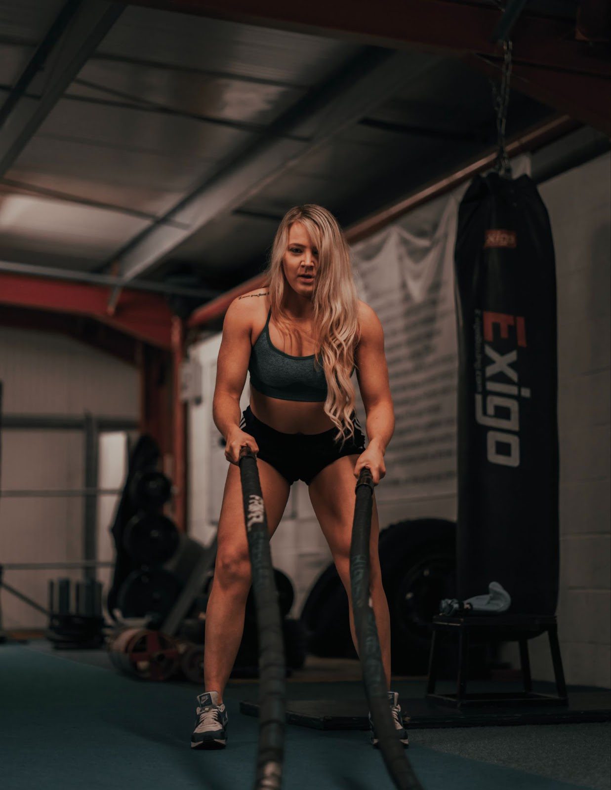 A muscular blonde woman in shorts and a sports bra holds weight ropes in a squat position at the gym.
