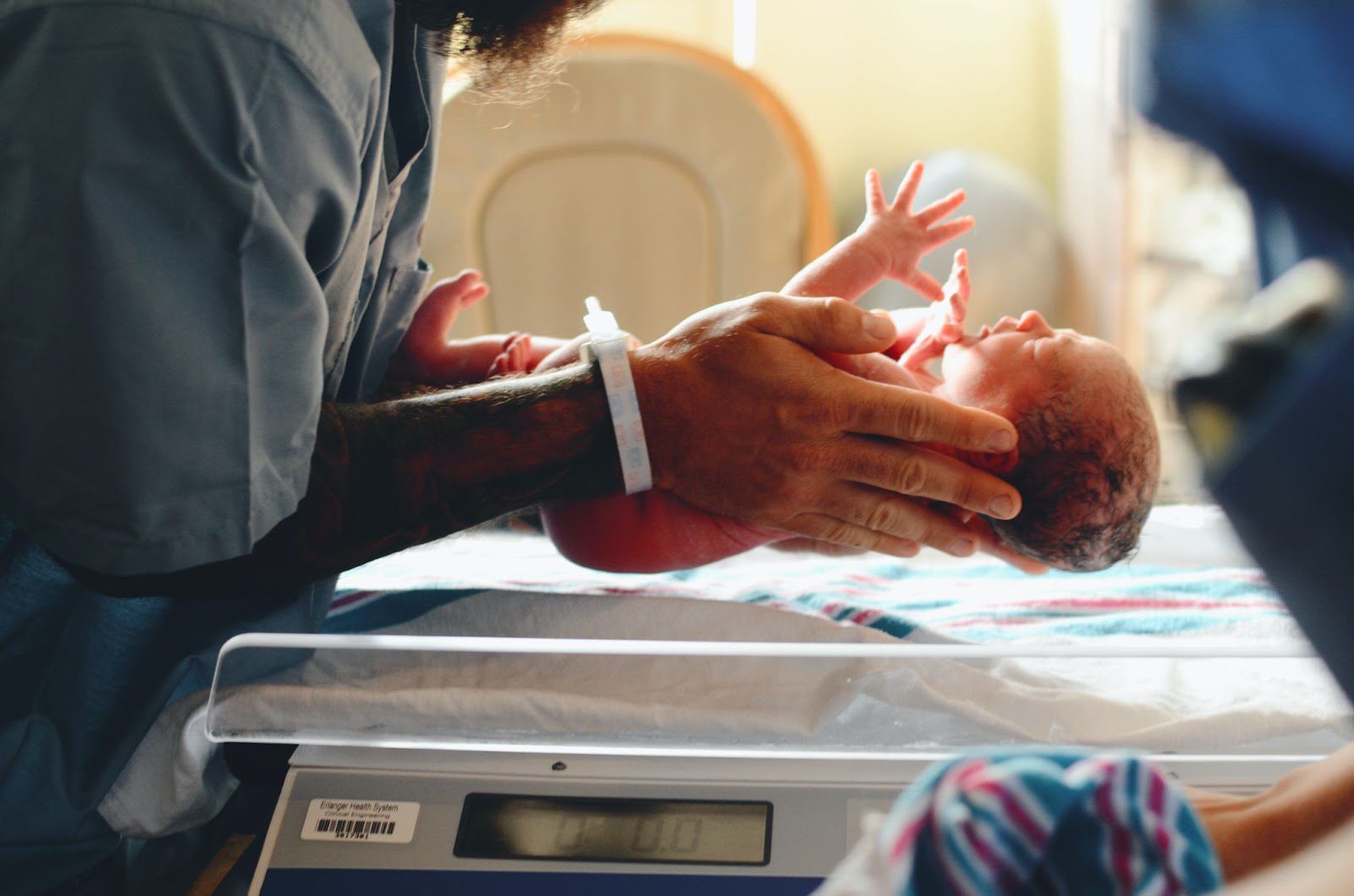 Newborn baby is held over a scale in the hands of a man with a tattooed arm and blue hospital scrubs.